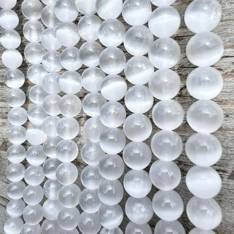 Selenite gemstone strands laid on a pieces of wood in outdoor and photographed under natural light. These beads are semi transparent with some white hues. Selenite beads have chatoyancy effect.