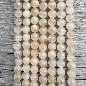 Może przedstawiać: Zbliżenie na sznury koralików z jasnożółtego cytrynu. Okrągłe koraliki są półprzezroczyste i nawleczone na cienką, czerwonopomarańczową nić. Tło stanowi zwietrzała, szara powierzchnia drewniana.