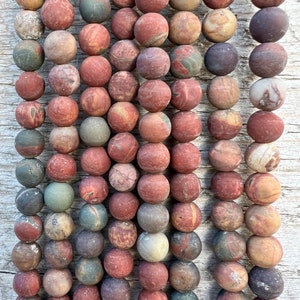 Puede incluir: Primer plano de una colección de cuentas redondas de piedra natural en varios tonos de rojo, marrón, gris y verde. Las cuentas están dispuestas en filas verticales, mostrando sus texturas y variaciones de color únicas. Ideales para la creación de joyas.