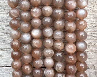 May include: Strands of round, polished moonstone beads in shades of peach, tan, and gray. The beads have a smooth, glossy finish and are strung together, creating a beautiful, natural gemstone jewelry component.
