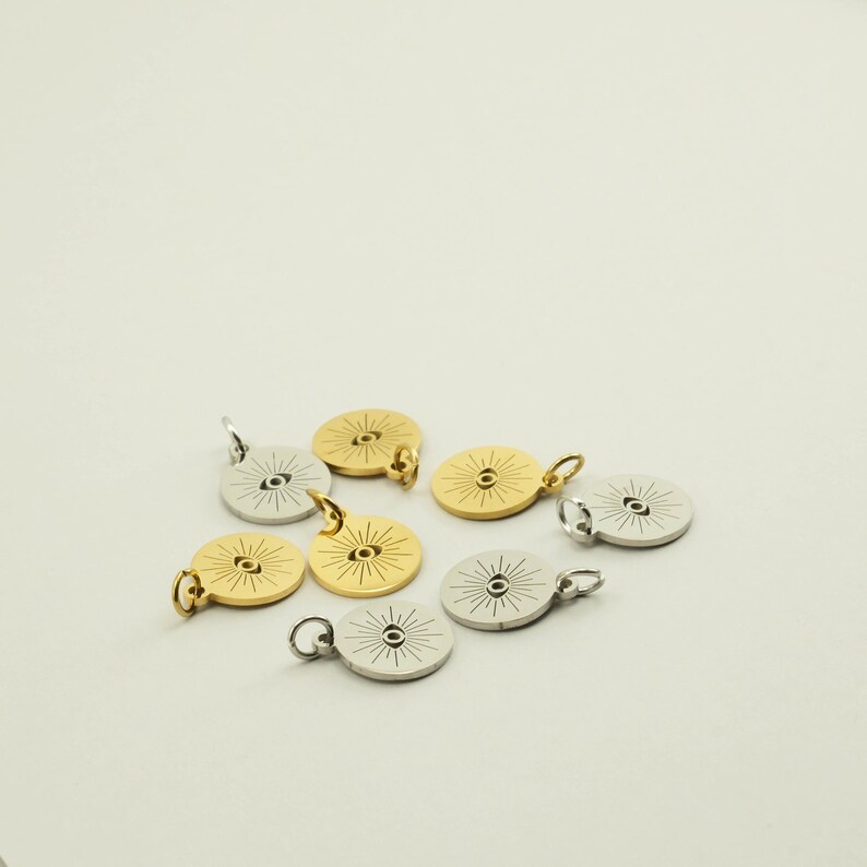May include: A collection of gold and silver-colored round pendants. Each pendant features an eye design with radiating lines. The pendants have small loops for attaching to a chain or other jewelry components. The pendants are scattered on a white surface.