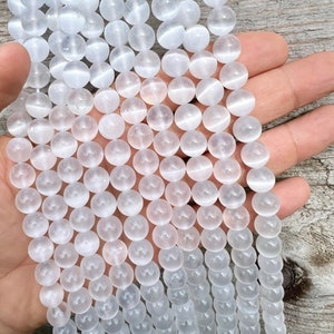 Selenite gemstone 10mm beads strands held in hand on a pieces of wood in outdoor and photographed under natural light. These beads are semi transparent with some white hues. Selenite beads have chatoyancy effect.