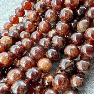 Può includere: Una collana di perle rotonde di granato marrone e rosso. Le perle sono lisce e lucide, con un leggero bagliore.