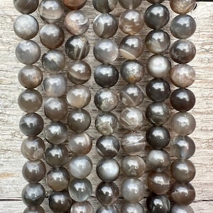 Può includere: Fili di perline rotonde e lucide in varie tonalità di grigio, marrone e argento. Le perline hanno una superficie liscia e riflettente e sono infilate insieme, creando un motivo strutturato e organico. Ideali per la creazione di gioielli.