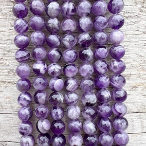 Könnte beinhalten: Eine Reihe runder, violetter Amethystperlen mit weißen Streifen. Die Perlen sind glatt und poliert und auf einer Holzoberfläche in einer Reihe angeordnet.