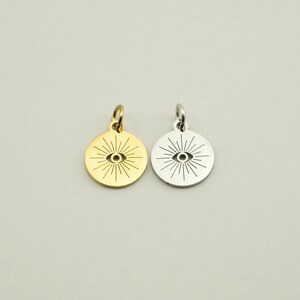 May include: Two round pendants, one gold-toned and one silver-toned, each featuring an eye design with radiating lines. The pendants have a small loop at the top for attaching to a chain or bracelet. The background is a solid, neutral color.