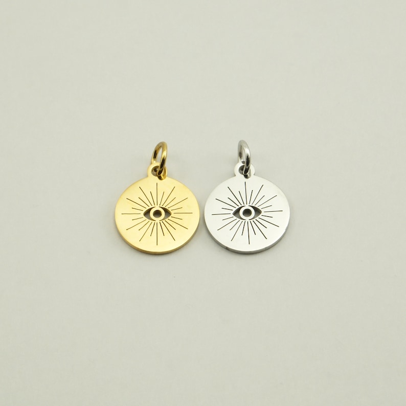 May include: Two round pendants, one gold-toned and one silver-toned, each featuring an eye design with radiating lines. The pendants have a small loop at the top for attaching to a chain or bracelet. The background is a solid, neutral color.
