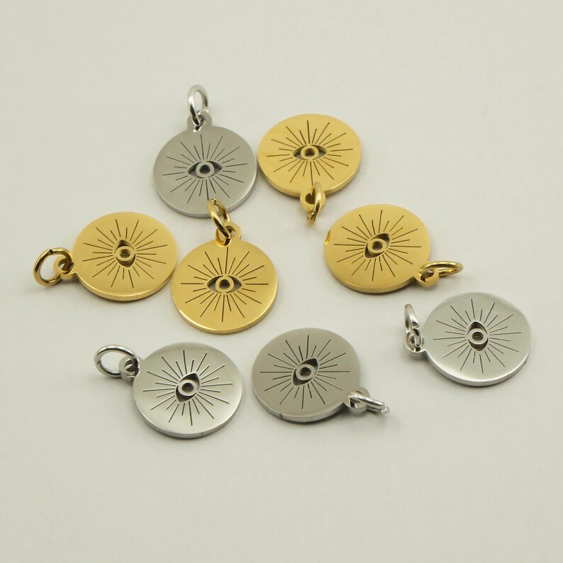 May include: A collection of round pendants with an eye design. The pendants are in gold and silver tones, with a sunburst pattern radiating from the eye. Each pendant has a small loop for attaching to a chain or bracelet.