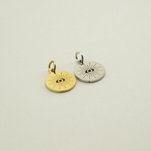 May include: Two round pendants, one gold-colored and one silver-colored, each featuring an eye design with radiating lines. The pendants have a small loop at the top for a chain. The background is a solid, neutral color.