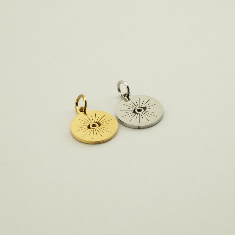 May include: Two round pendants, one gold-colored and one silver-colored, each featuring an eye design with radiating lines. The pendants have a small loop at the top for a chain. The background is a solid, neutral color.