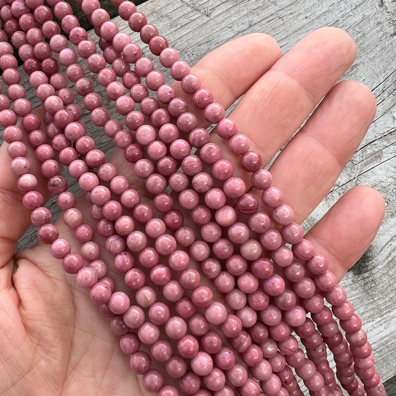 Rhodonite beads strands held in hand on a bright wooden board in outdoor under natural light. These are round 6mm beads with 1mm hole in middle for threading. they have a very delicate vein on them.