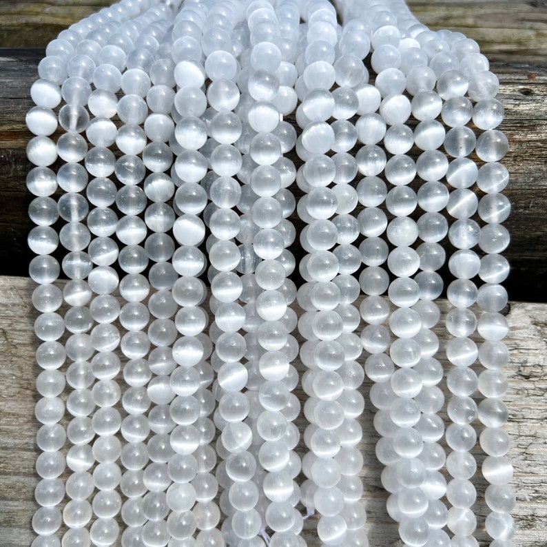 Selenite gemstone strands laid on a pieces of wood in outdoor and photographed under natural light. These beads are semi transparent with some white hues. Selenite beads have chatoyancy effect.pictured from front