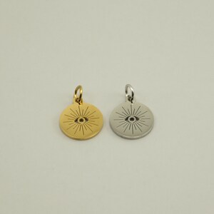 May include: Two round pendants, one gold-toned and one silver-toned, each featuring an eye design with radiating lines. The pendants have a small loop at the top for attaching to a chain or bracelet. The background is a solid, neutral color.