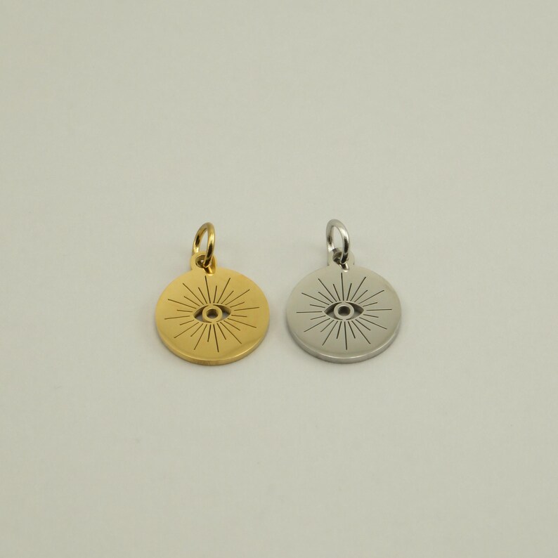 May include: Two round pendants, one gold-toned and one silver-toned, each featuring an eye design with radiating lines. The pendants have a small loop at the top for attaching to a chain or bracelet. The background is a solid, neutral color.