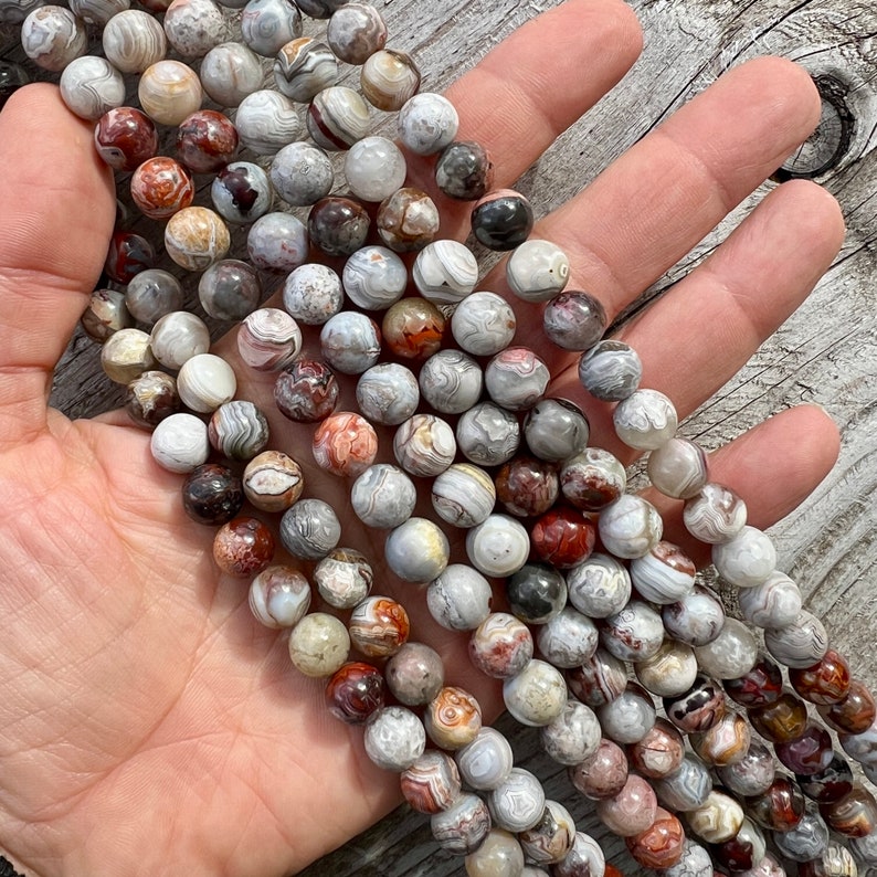 Laguna Lace Agate 10mm beads strands held in hand on a bright wood board in outdoors under natural daylight. these beads are white and gray and have lots of veins in various shapes and colors from red to brawn or pale blue