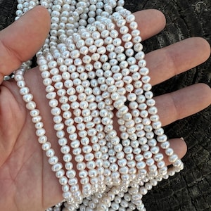 Könnte beinhalten: Eine Strähne weißer Süßwasserperlen, etwa 5-6 mm groß. Die Perlen sind rund und haben eine glatte, glänzende Oberfläche. Sie sind auf einem dünnen, weißen Faden aufgefädelt.