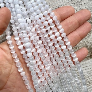 Selenite gemstone 6mm beads strands held in hand on a pieces of wood in outdoor and photographed under natural light. These beads are semi transparent with some white hues. Selenite beads have chatoyancy effect.