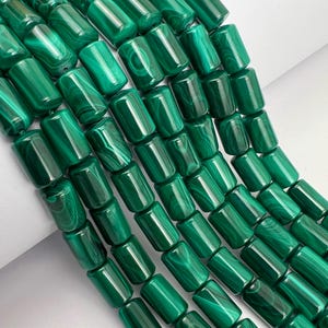 Pode incluir: Uma aproximação de um fio de contas de malaquite polidas. As contas são de cor verde-escura com padrões em espiral e de forma cilíndrica. As contas são enfiadas, criando uma exibição texturizada. As contas têm cerca de 2,5 cm de comprimento.