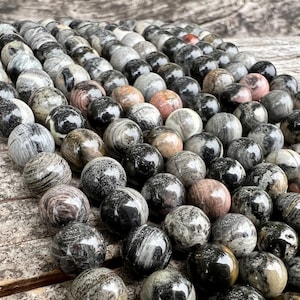 Könnte beinhalten: Eine Sammlung polierter Steinkugeln in verschiedenen Farben, darunter Schwarz, Grau, Weiß und Rosa. Die Kugeln haben ein marmoriertes Aussehen und sind eng aneinandergereiht, wodurch eine strukturierte Oberfläche entsteht. Die Kugeln liegen auf einer Holzoberfläche.