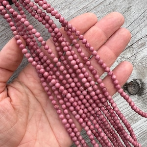 Rhodonite beads strands held in hand on a bright wooden board in outdoor under natural light. These are round 4mm beads with 1mm hole in middle for threading. they have a very delicate vein on them.