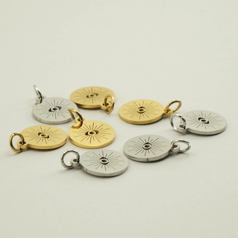 May include: A collection of round pendants in gold and silver tones. Each pendant features an eye design with radiating lines. The pendants have a small loop for attaching to a chain or bracelet. The pendants are displayed on a white surface.