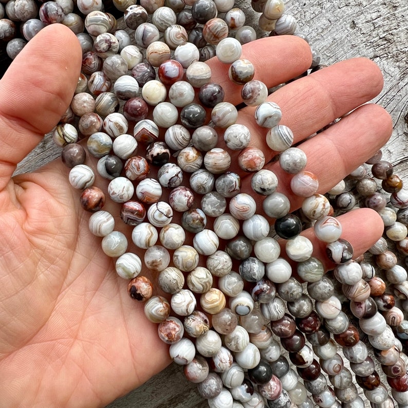 Laguna Lace Agate 8mm beads strands held in hand on a bright wood board in outdoors under natural daylight. these beads are white and gray and have lots of veins in various shapes and colors from red to brawn or pale blue