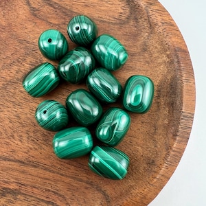 Könnte beinhalten: Eine Sammlung polierter, grüner Malachitperlen mit wirbelnden Mustern. Die oval geformten Perlen haben eine glänzende Oberfläche. Sie werden auf einer Holzoberfläche präsentiert, um ihre natürliche Schönheit hervorzuheben. Ideal für die Schmuckherstellung.