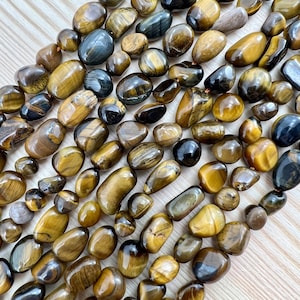 Könnte beinhalten: Eine Strähne aus Tigerauge-Edelsteinen in getrommelter, kieseliger Form. Die Perlen sind eine Mischung aus goldbraun und schwarz mit einem glänzenden Finish.