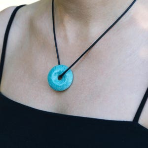 Può includere: Una collana con un ciondolo a forma di ciambella turchese. Il ciondolo è sospeso a un cordino nero. La collana è indossata su un top nero.