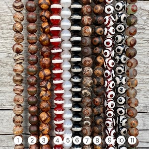Op de afbeelding: Een verzameling kralenstrengen met verschillende ontwerpen en kleuren. De kralen zijn bruin, rood, wit en zwart, met patronen zoals strepen, stippen en oogachtige motieven. De strengen zijn genummerd van 1 tot en met 11.