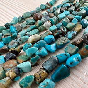 Könnte beinhalten: Stränge aus türkisfarbenen Edelsteinperlen in verschiedenen Blau-, Grün- und Brauntönen. Die Perlen sind unregelmäßig geformt und groß, auf einer blauen Schnur aufgereiht. Die Natursteinperlen haben eine polierte Oberfläche.