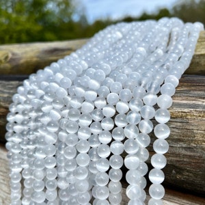 Selenite gemstone strands laid on a pieces of wood in outdoor and photographed under natural light. These beads are semi transparent with some white hues. Selenite beads have chatoyancy effect.pictured from side