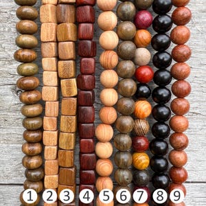 Op de afbeelding: Negen strengen houten kralen in verschillende kleuren en patronen. De kralen zijn rond en vierkant, sommige hebben een natuurlijke houten afwerking terwijl andere zijn gebeitst. De kralen zijn genummerd van 1 tot en met 9.