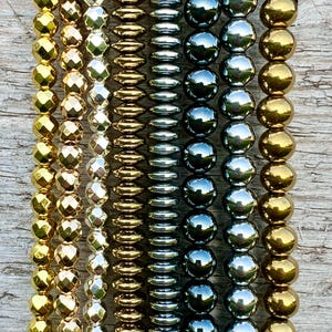 Può includere: Primo piano di dieci fili di perline. Le perline sono realizzate con vari materiali, tra cui oro, argento e nero. Le perline sono tutte di forme e dimensioni diverse.