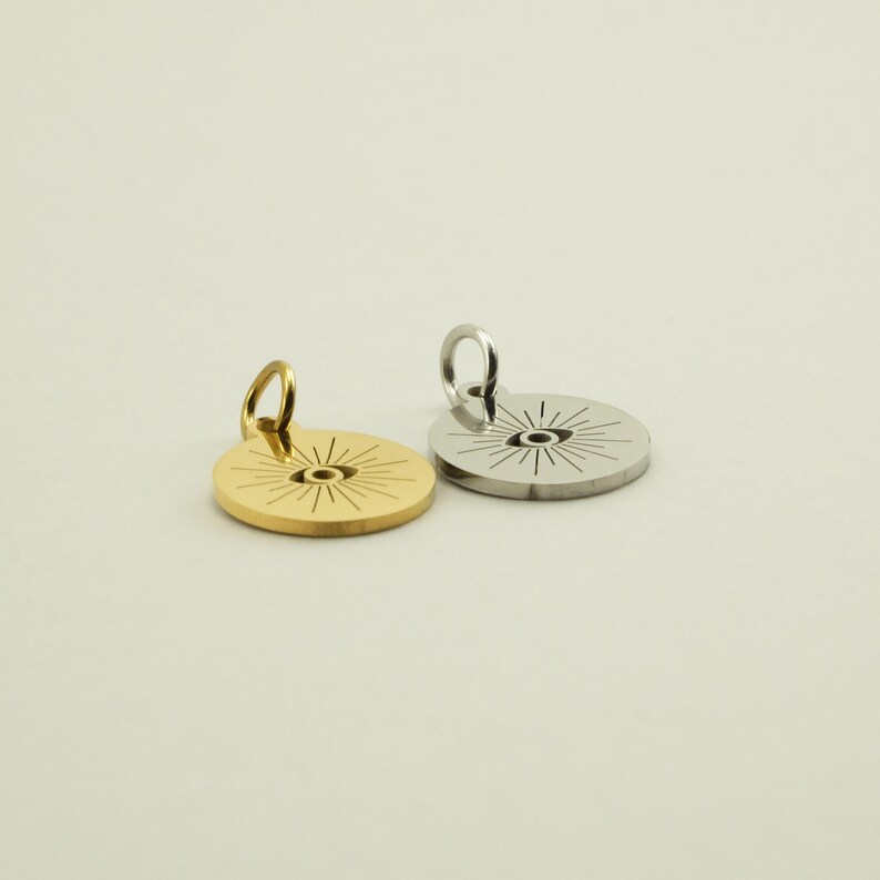 May include: Two round pendants with a sunburst design. One pendant is gold-colored, and the other is silver-colored. Each pendant has a small loop at the top for attaching to a chain or bracelet. The pendants are on a light background.