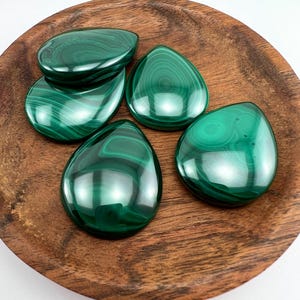Peut inclure: Cinq pierres précieuses de malachite polies, en forme de goutte d'eau, sont présentées sur une assiette en bois. Les pierres sont d'un vert profond avec des motifs tourbillonnants. L'assiette en bois présente un grain de bois naturel.