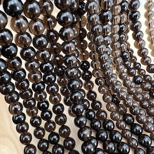 Puede incluir: Una hebra de cuentas de cuarzo ahumado marrón oscuro. Las cuentas son redondas y lisas, y tienen una apariencia translúcida.