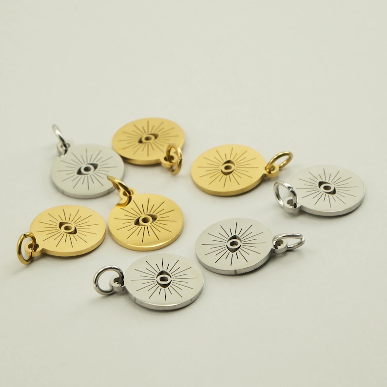 May include: A collection of gold and silver-colored round pendants. Each pendant features a central eye design with radiating lines. The pendants have small loops for attaching to a chain or bracelet. The pendants are likely for jewelry making.