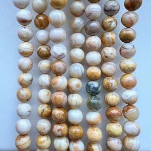 May include: A collection of round, polished gemstone beads in cream, brown, and tan hues. The beads display unique patterns and textures, offering a natural, earthy appearance. The beads are strung together, suitable for jewellery making.