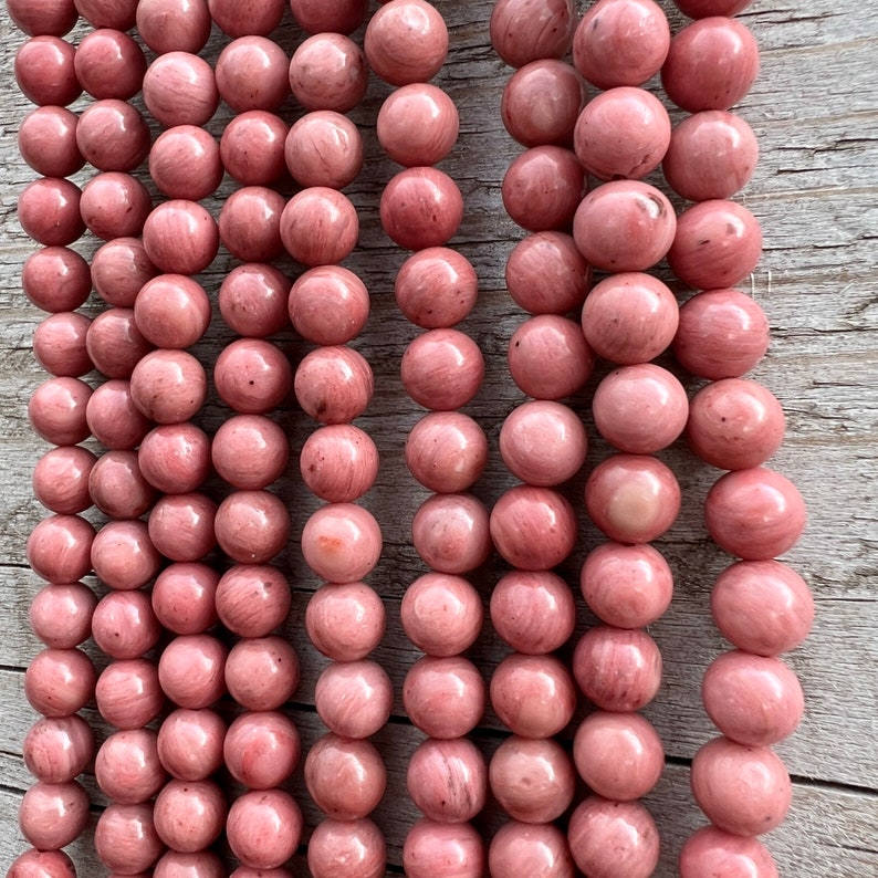 Rhodonite beads strands laid on a bright wooden board in outdoor under natural light. These are round 8mm beads with 1mm hole in middle for threading. they have a very delicate vein on them.