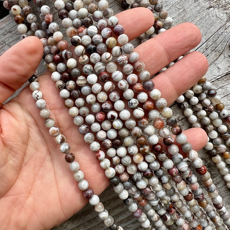 Laguna Lace Agate 6mm beads strands held in hand on a bright wood board in outdoors under natural daylight. these beads are white and gray and have lots of veins in various shapes and colors from red to brawn or pale blue