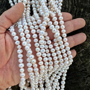 Könnte beinhalten: Eine Strähne weißer Süßwasserperlen, etwa 6-7 mm groß. Die Perlen sind rund und glatt, mit einem hohen Glanz. Sie sind auf einem weißen Faden aufgefädelt.