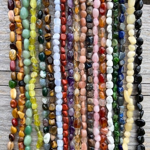 Op de afbeelding: Een verzameling strengen edelsteen kralen in verschillende kleuren. De kralen zijn in tinten bruin, groen, geel, rood, paars, roze, wit, blauw en zwart. De kralen hebben verschillende vormen en maten, geregen voor sieraden.