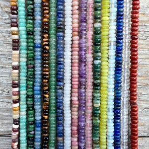 May include: A collection of colorful gemstone beads in various shades, including green, blue, pink, and red. The beads are strung together, creating a vibrant display of natural stone jewelry components. The beads are arranged vertically, showcasing their diverse colors and textures.