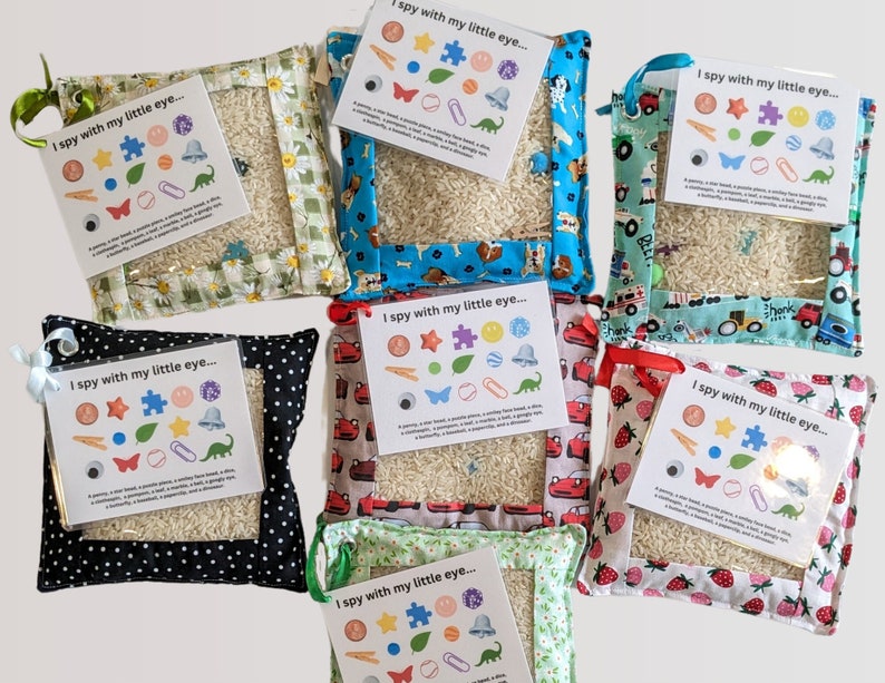 May include: Seven square fabric pouches filled with rice, each featuring a clear plastic window and an attached card titled I spy with my little eye. The pouches have various patterns including daisies, dogs, vehicles, polka dots, and strawberries.