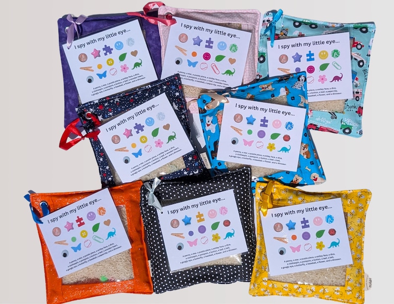 May include: A collection of eight square fabric pouches in various colors and patterns, each featuring a clear window and a card labeled I spy with my little eye. The cards display colorful icons of small objects like a penny, star, puzzle piece, and dinosaur, which are hidden inside the pouches.