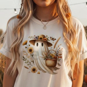 Op de afbeelding: Crèmekleurig T-shirt met een grillige geest met een strohoed versierd met zonnebloemen en andere bloemen. De geest houdt een bloemenmand vast, met vlinders en tarwearen eromheen. Het ontwerp combineert herfst- en Halloween-thema's.