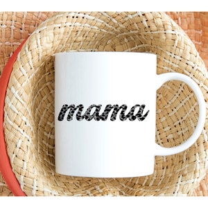 以下が含まれることがあります： 白のセラミックマグカップ。黒と白の模様のフォントで「mama」の文字が書かれています。マグカップは、赤いバンドが付いた麦わら帽子の上に置かれています。