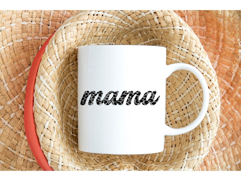 以下が含まれることがあります： 白のセラミックマグカップ。黒と白の模様のフォントで「mama」の文字が書かれています。マグカップは、赤いバンドが付いた麦わら帽子の上に置かれています。