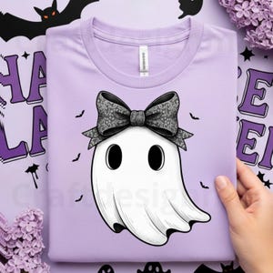 Op de afbeelding: Een lichtpaars t-shirt met een witte geest met een patroonstrik. De geest heeft grote zwarte ogen en is omringd door kleine zwarte vleermuizen. Het shirt is opgevouwen en het ontwerp is gecentreerd op de voorkant.